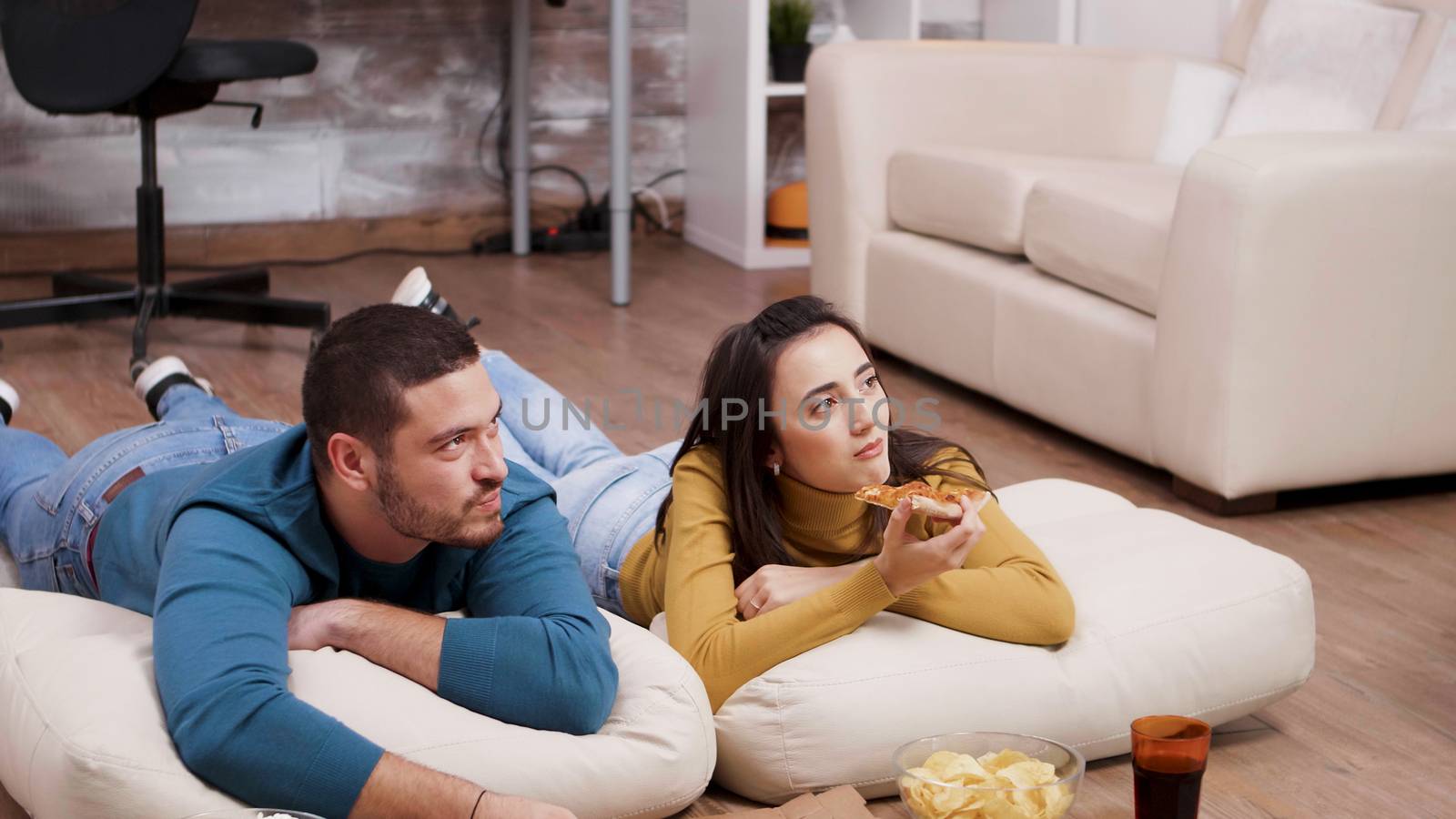 Young man and woman lying on the floor watching tv and eating pizza with the back in the background.