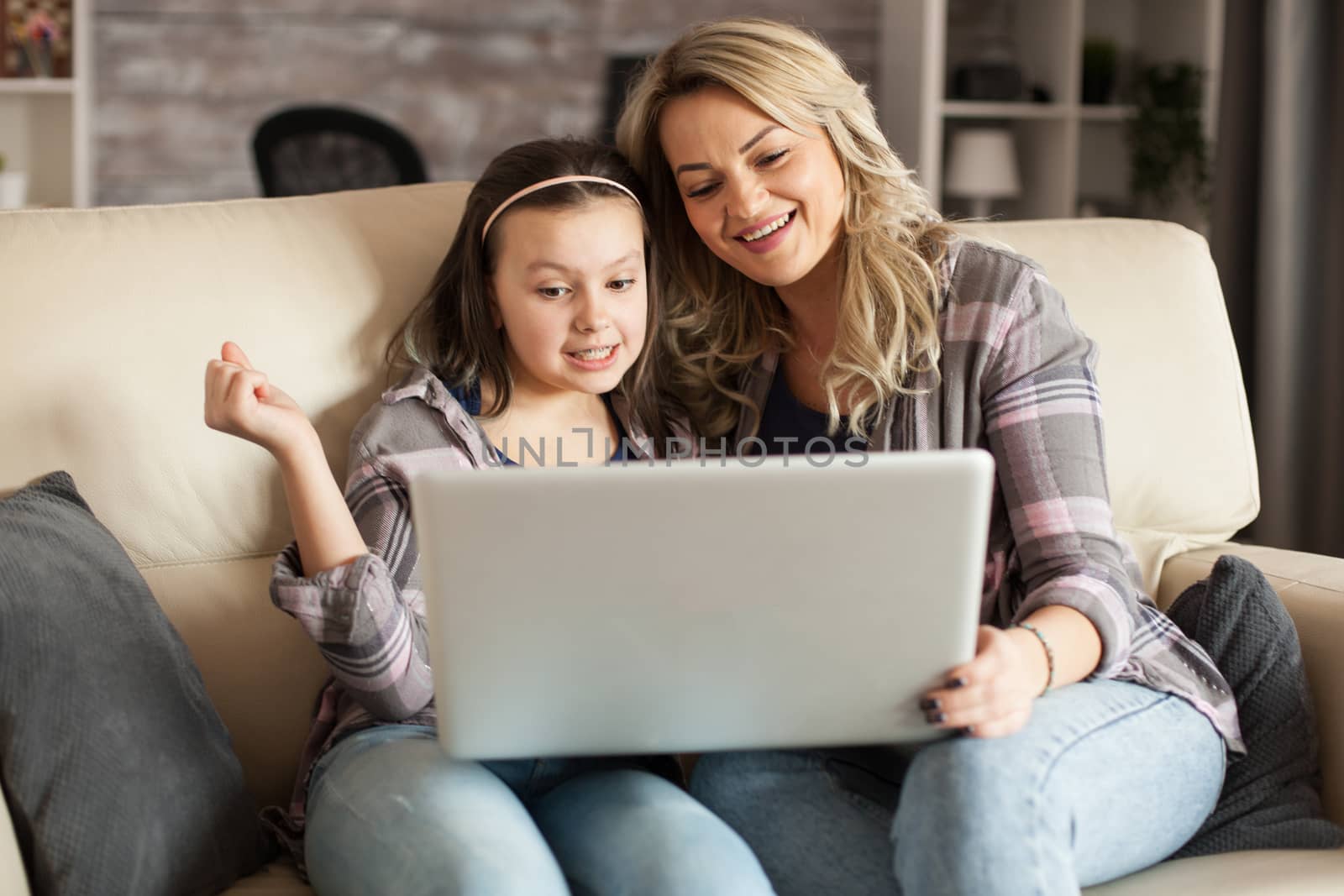 Cheerful young mother and her daughter smiling while surfing on laptop sitting on the couch in living room.