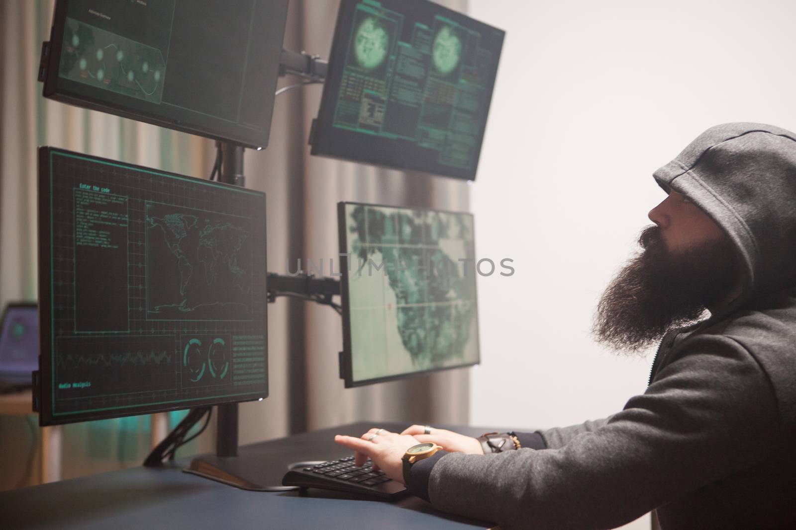 Bearded cyberterrorist wearing a hoodie writing a dangerous virus. Hacker using computer with multiple screens.