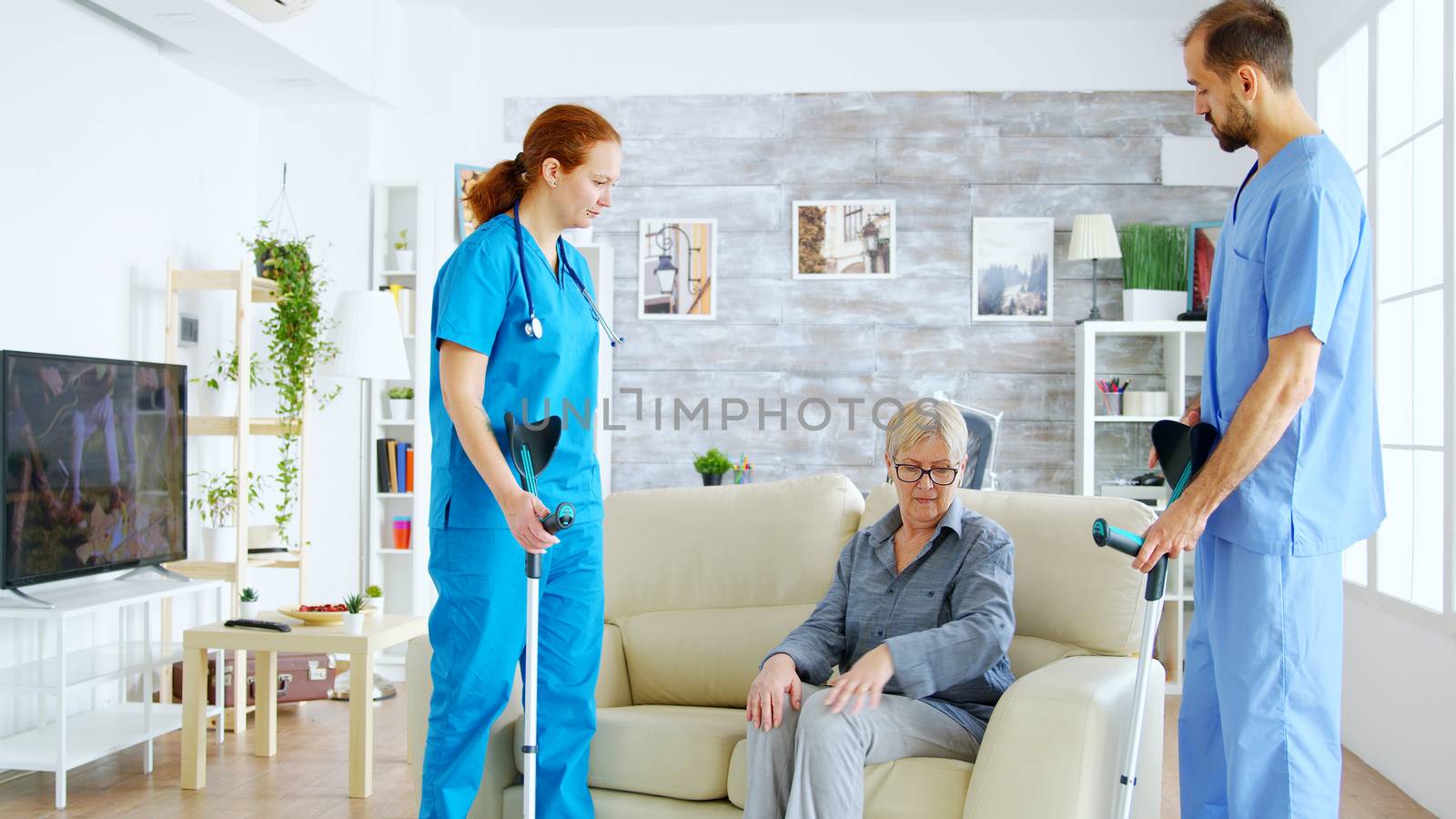 Female doctor and her assistant helping old woman with crutches to stand up from the couch by DCStudio