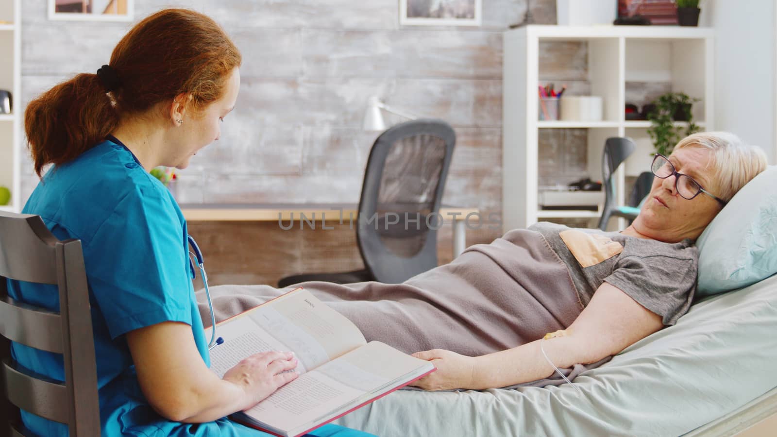 Revealing shot of an old lady lying in hospital bed while a nurse is reading a book to her. Caregiver and social worker