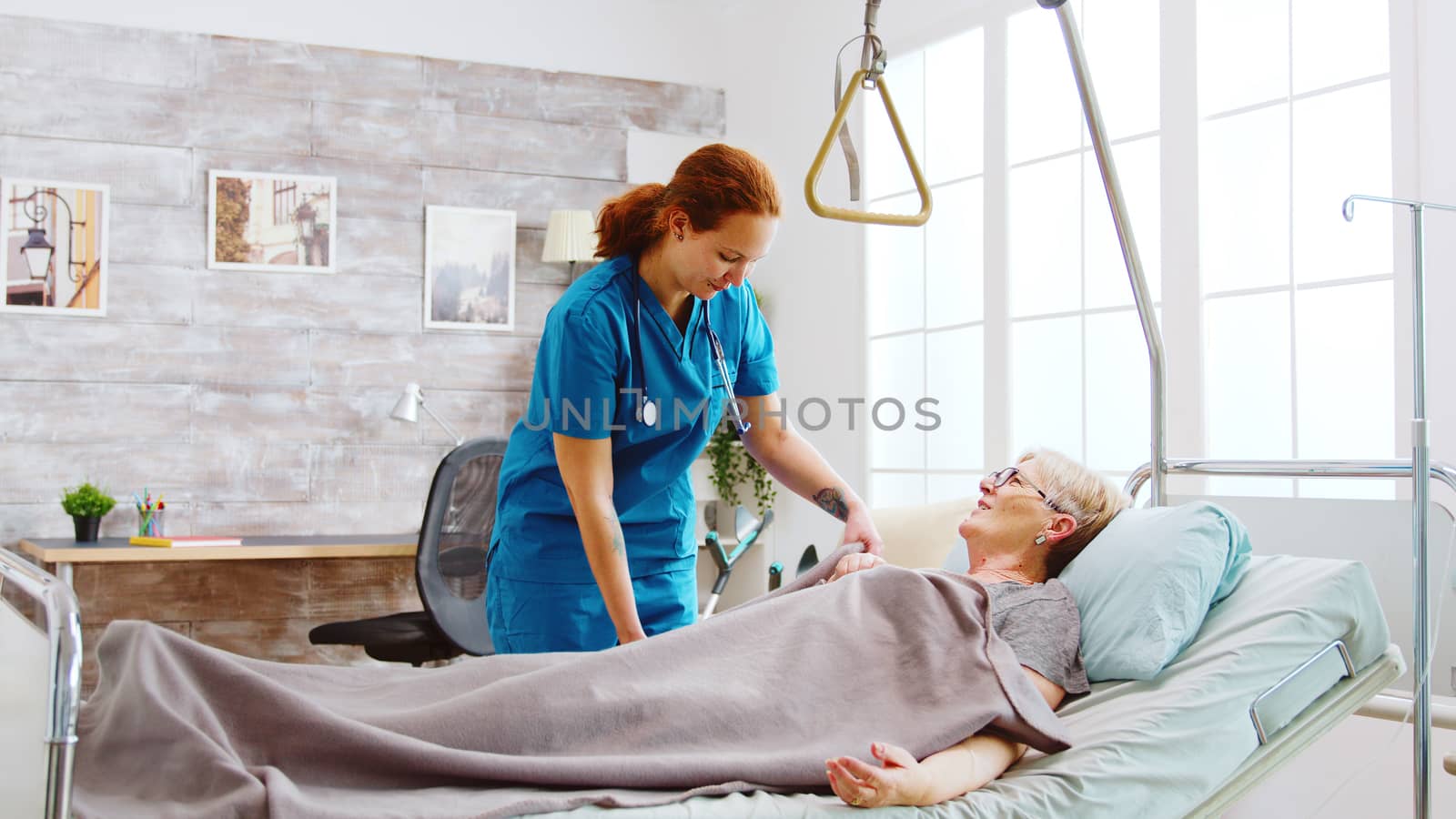 Female nurse helping an old disabled and retired woman to go to bed. The caregiver is covering her with a blanket.