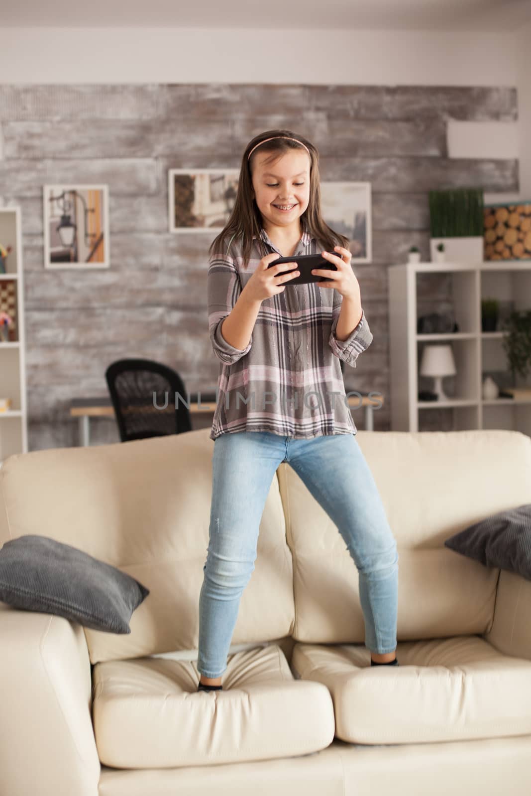 Little girl playing games on smartphones jumping on the couch in living room.