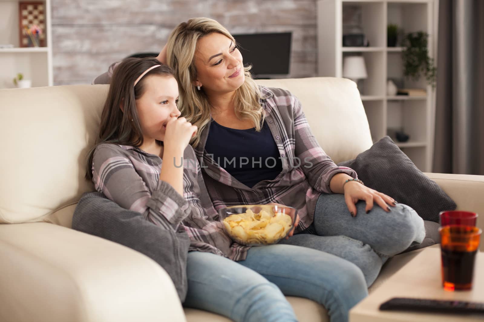 Cheerful daughter and her young mother relaxing watching a film on tv. Snacks and soda.