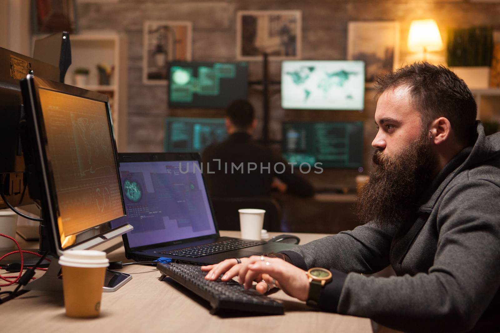 Focused bearded hacker after drinking a lot of coffee to stay awake. Young cyber criminal in the background.