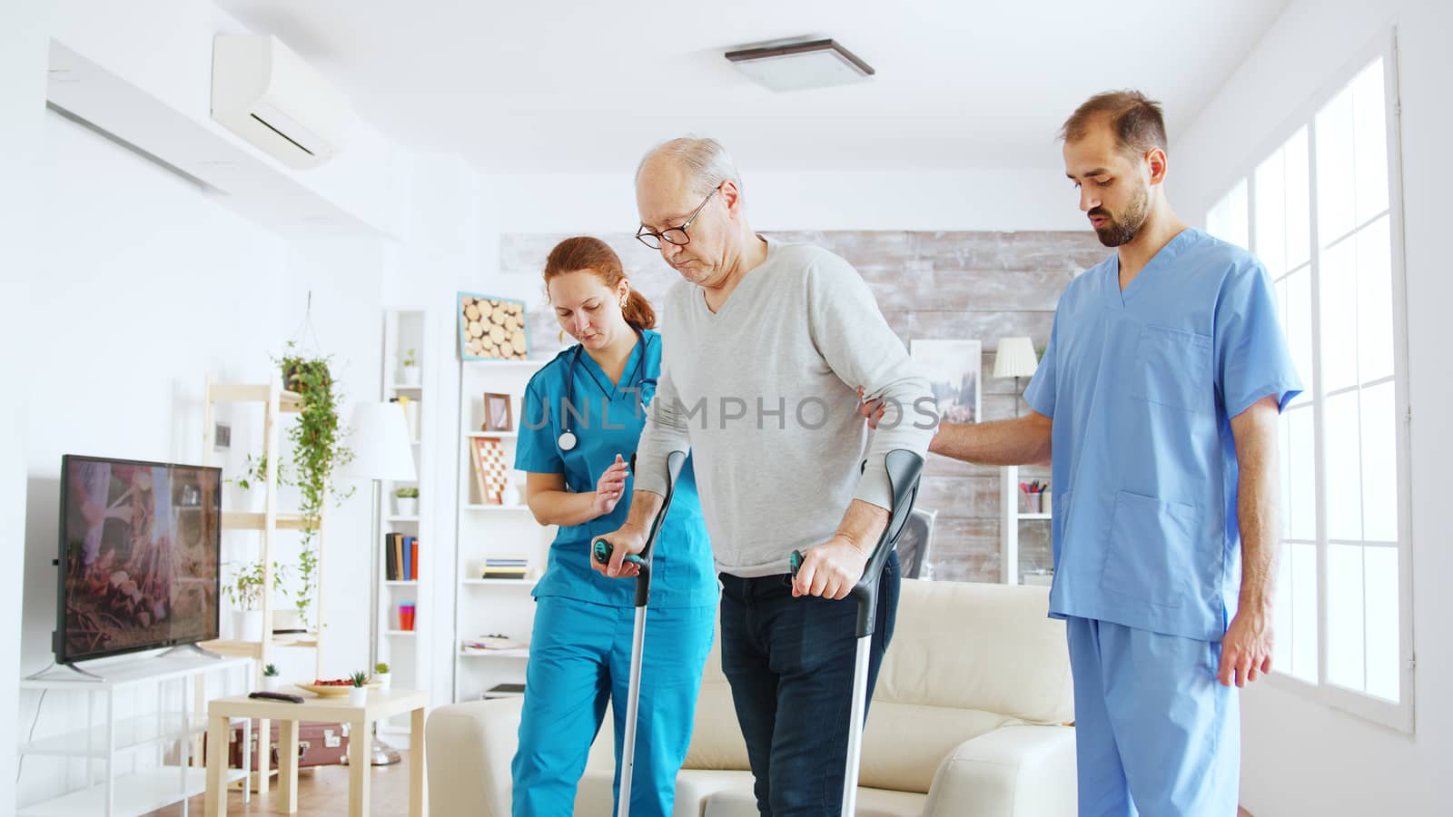 Team of nurses or social workers helping an old disabled man to walk with his crutches out of the nursing home room by DCStudio