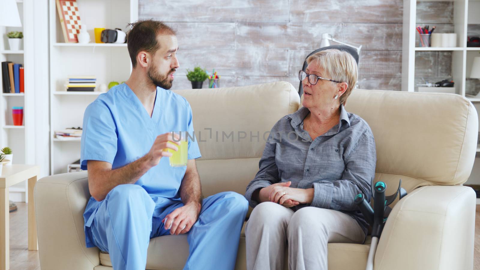 Male nurse sitting on couch with senior woman giving her medical treatment in nursing home by DCStudio