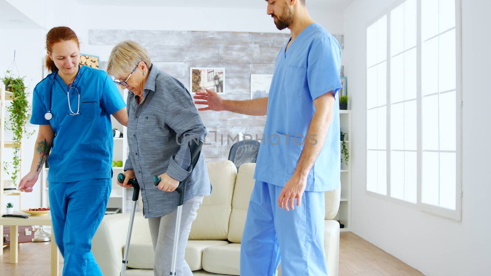 Female doctor and her assistant helping old woman with crutches to stand up from the couch and take a walk