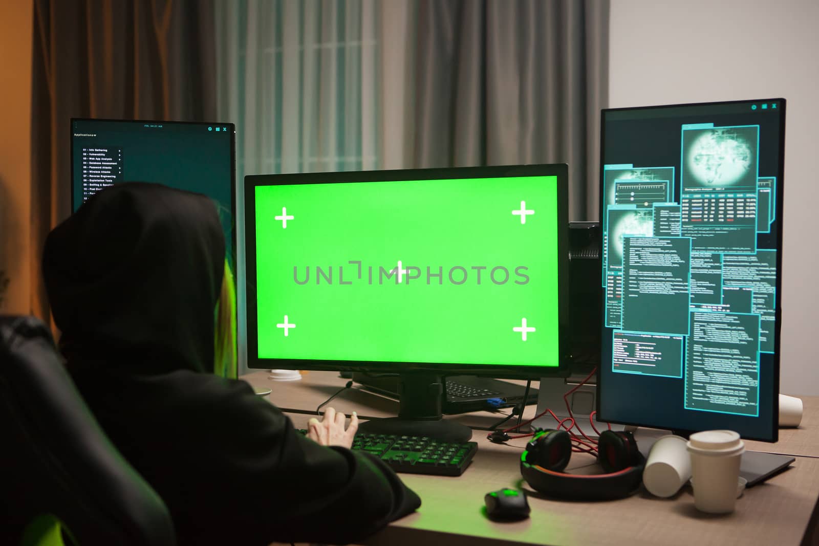 Dangerous hooded female hacker in front of computer with green screen.