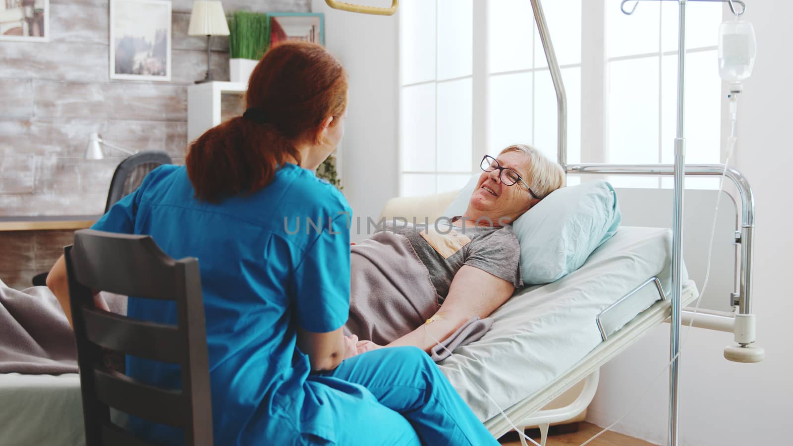 In bright retirement home old lady lying in hospital bed talks with a female caregiver.