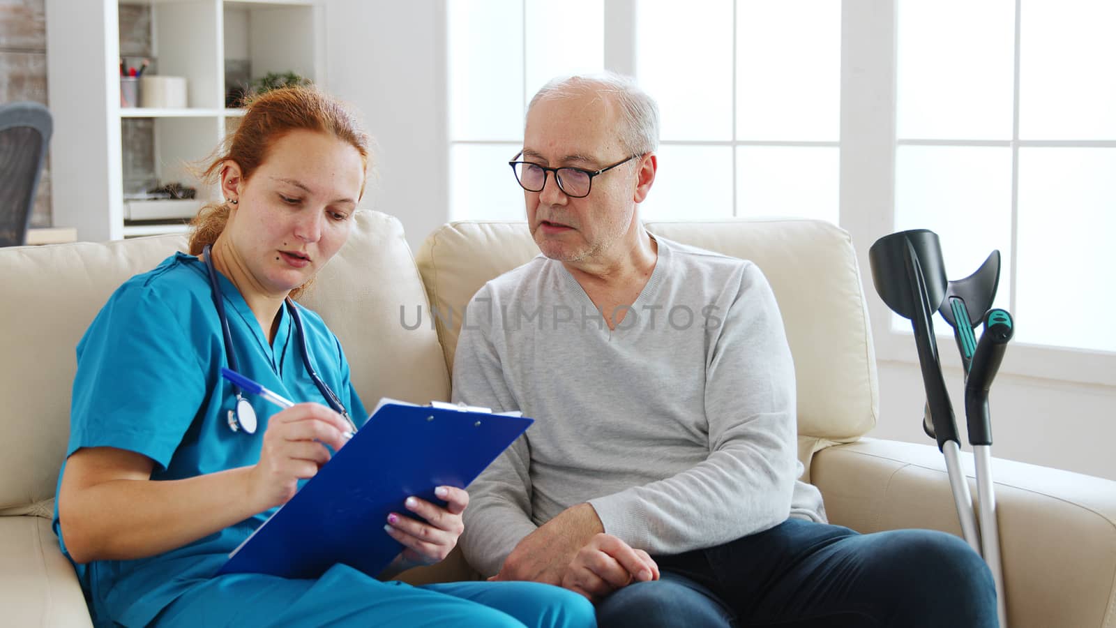 Female healthcare assistant taking notes about an old man health in cozy nursing home. They chat sitting on the sofa