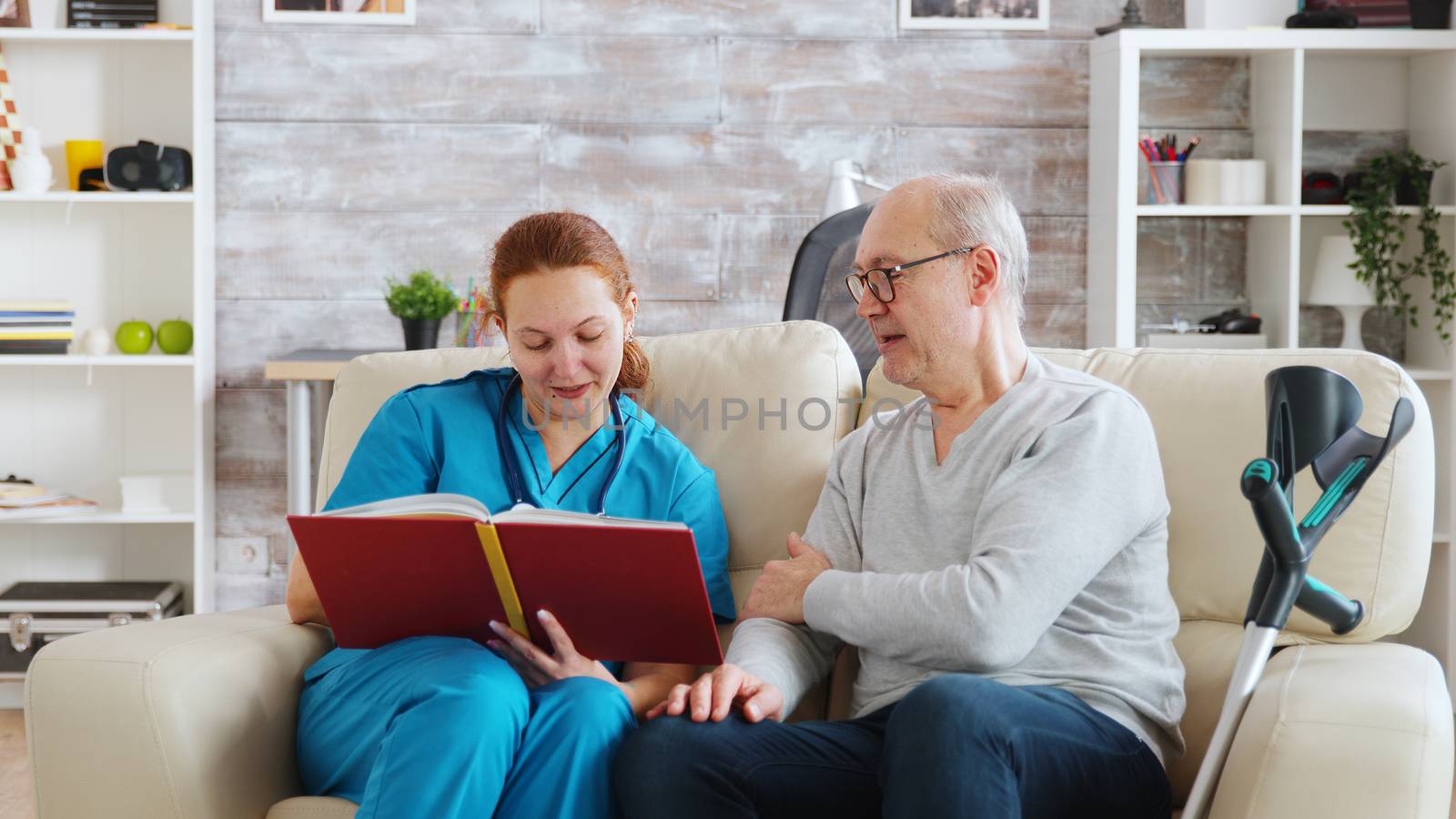 Social worker reading to an old and disabled man sitting on a sofa in bright nursing home