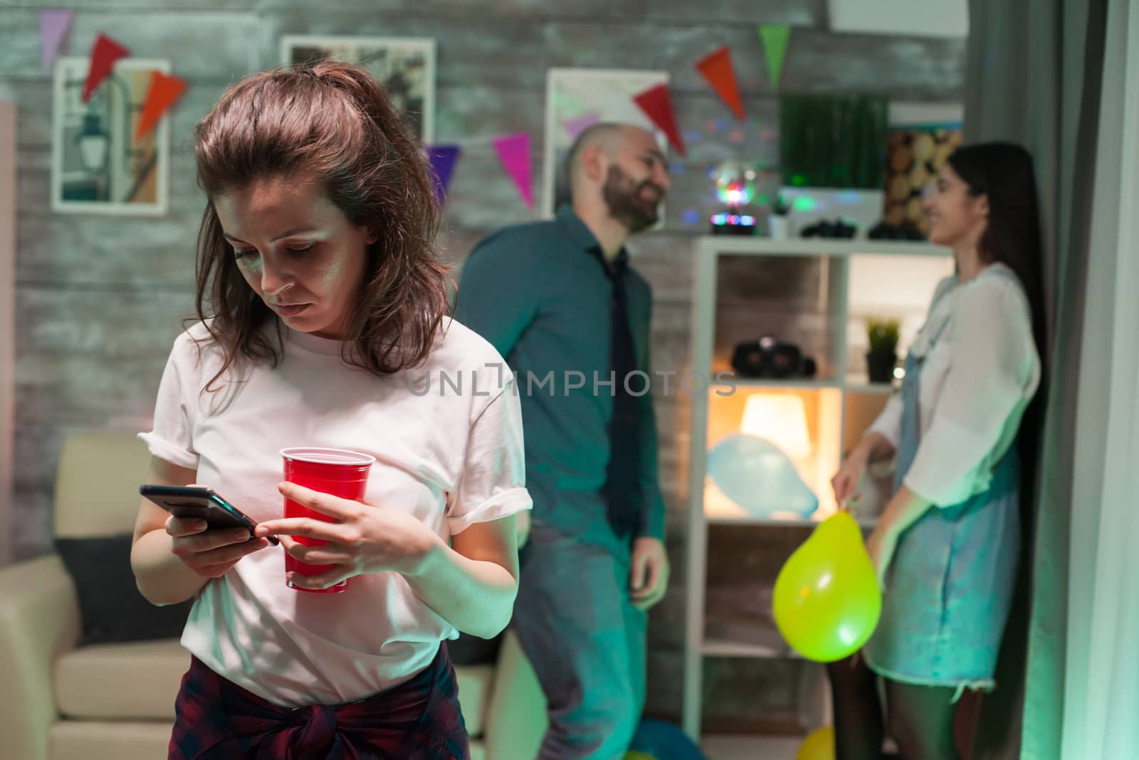 Young woman using her smartphone at a cheerful party. Man and woman talking in the background.