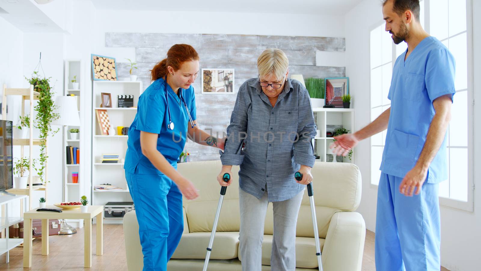 Senior woman with crutches getting help for female nurse and doctor to stand on her legs from the couch