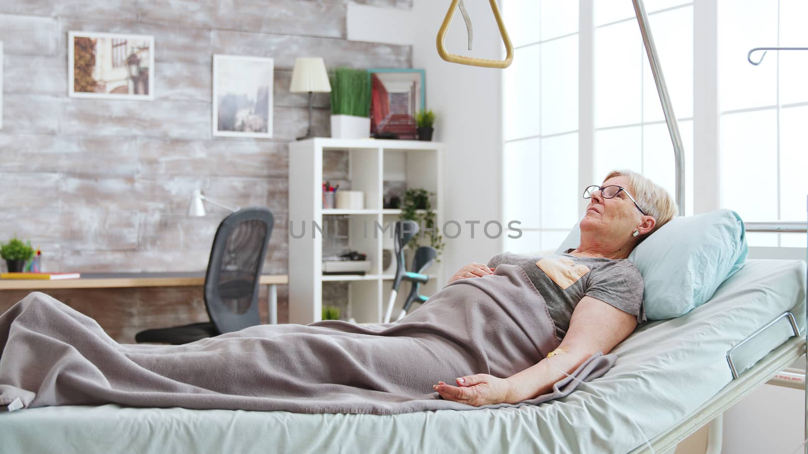 Disabled old lady lying in hospital bed in bright and cozy nursing home by DCStudio