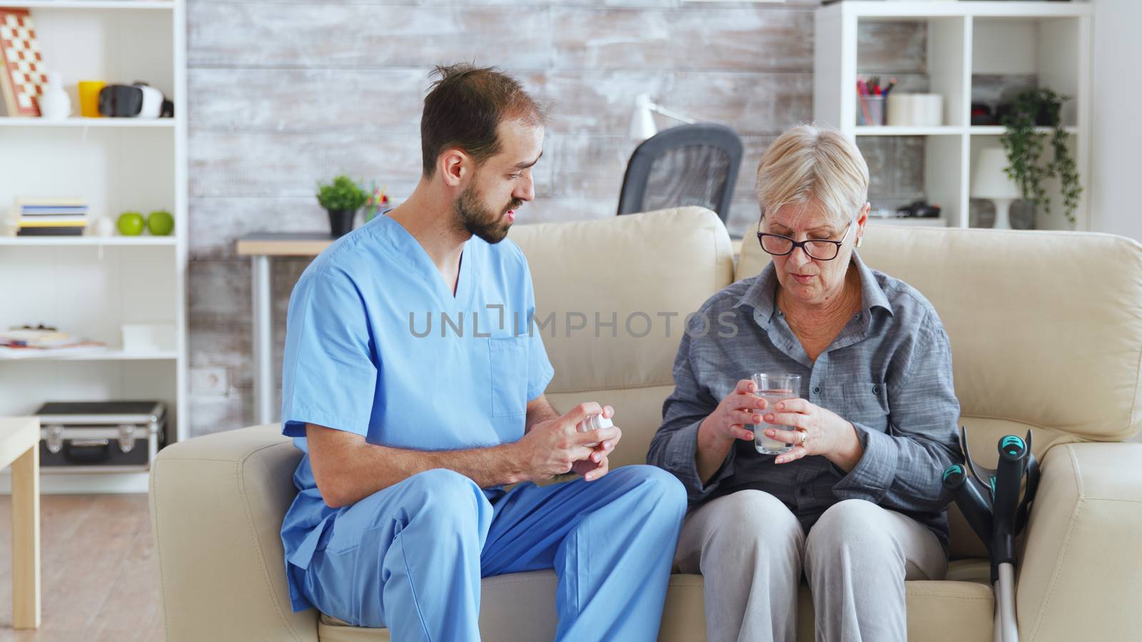 Male nurse sitting on couch with senior woman giving her medical treatment in nursing home.