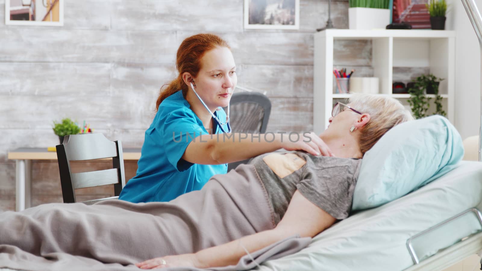 Female nurse listening to old lady heartbeats. She is lying in a hospital bed in a bright cozy nursing home