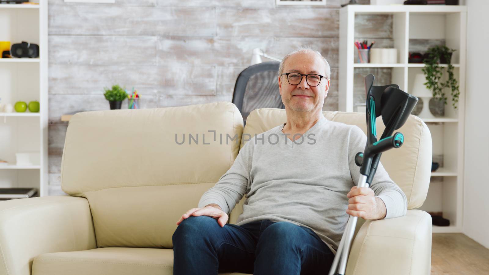 Portrait of elderly retired man smiling to the camera in cozy nursing home, he has crutches next to the sofa