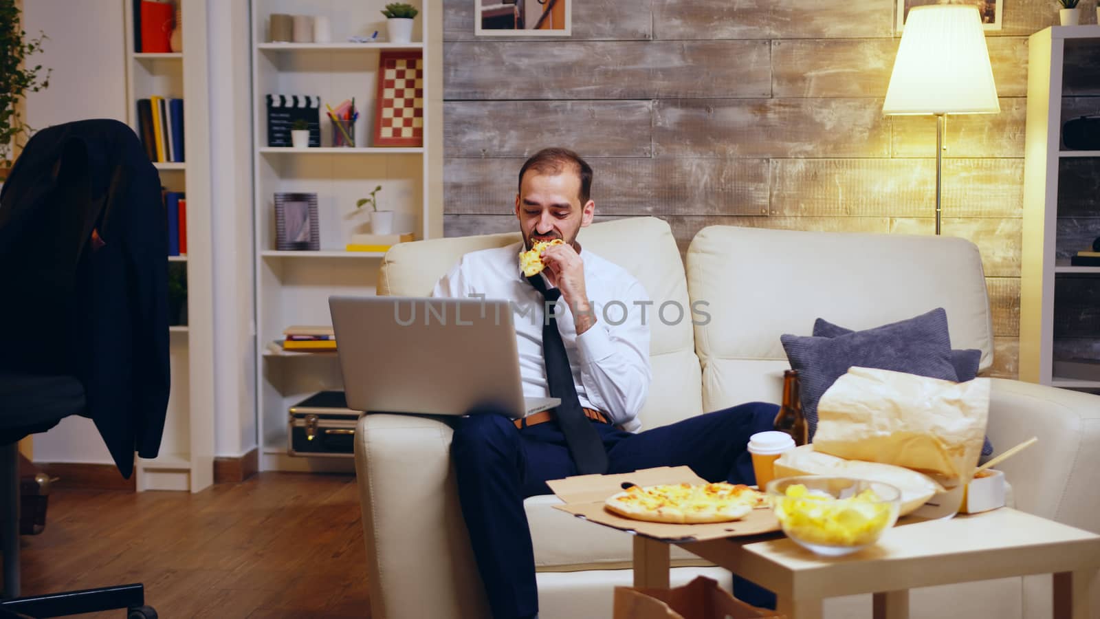 Man in business suit working late at night on the laptop in front of his TV, eating junk food.