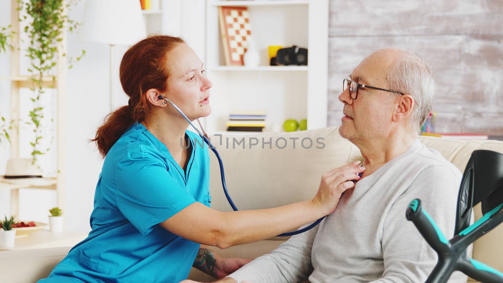Caucasian female nurse listening to elderly retired man heartbeat in bright and cozy nursing home. Social worker and caregiver