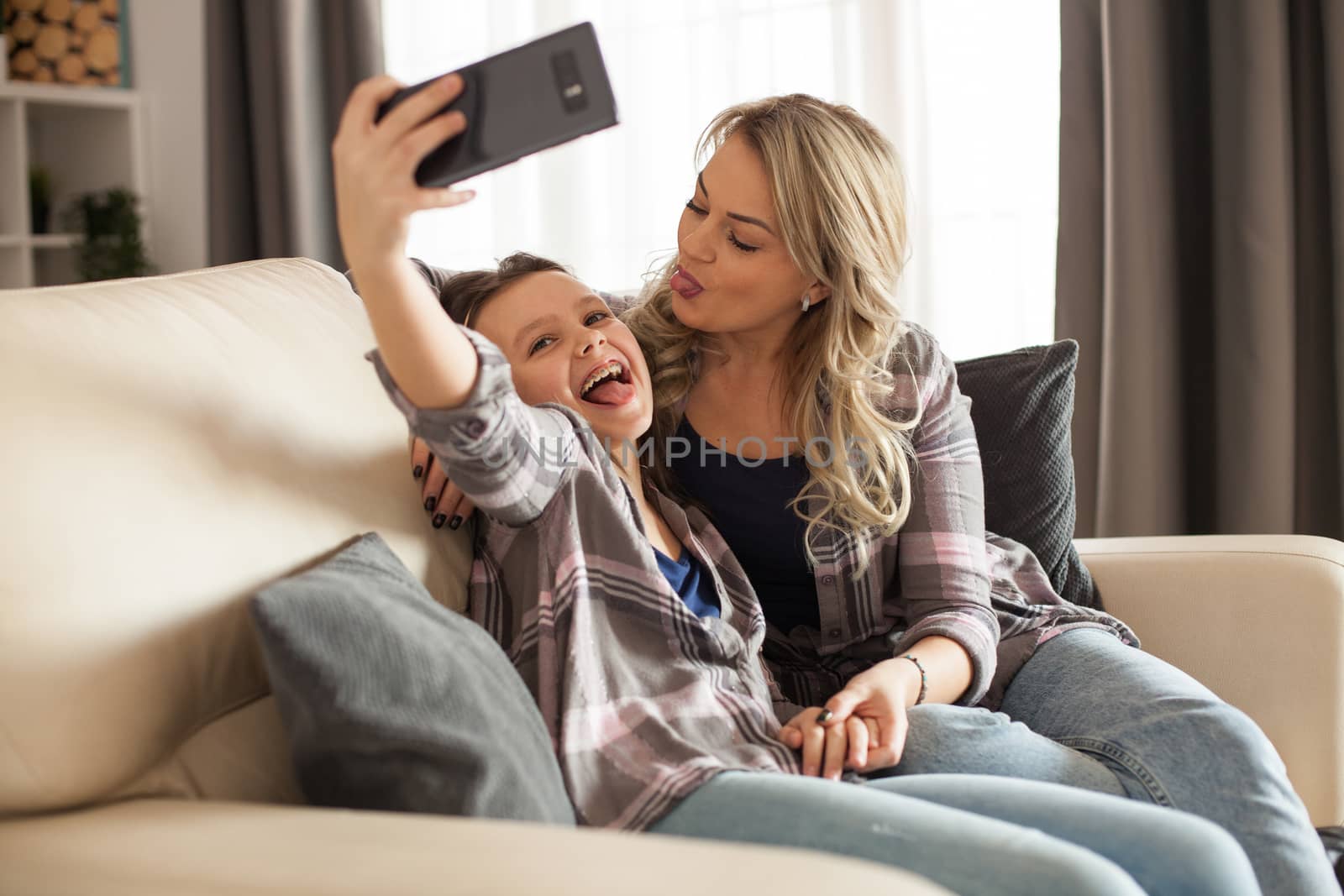 Cheerful mother and daughter sitting on the couch in living room taking a selfie together.