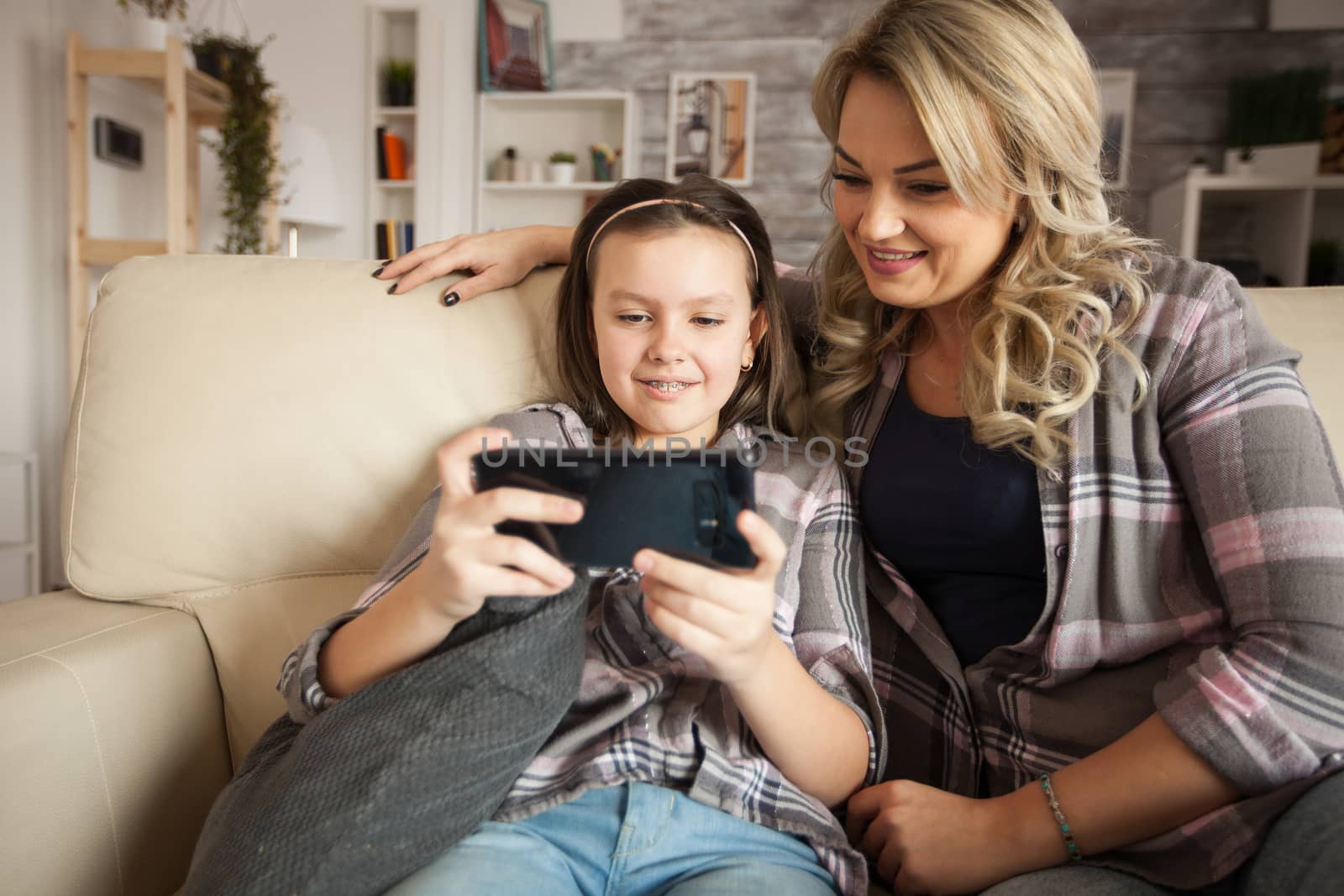 Modern mum and happy little girl sitting on the couch in living room using smartphone application.