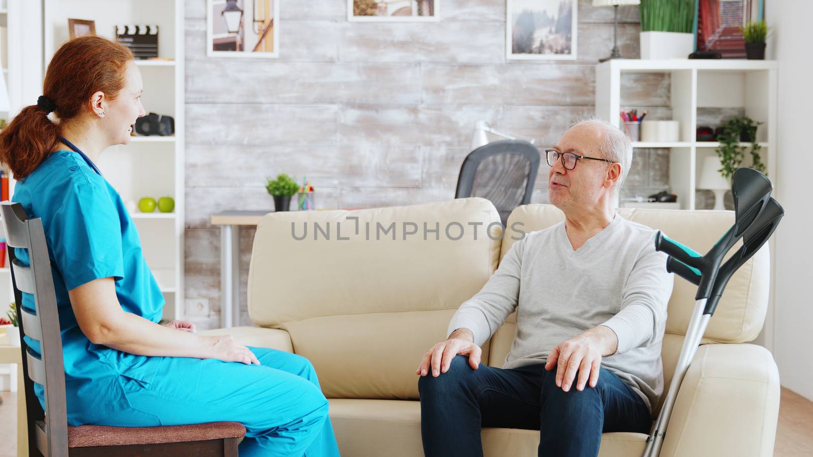 Elderly disabled man with crutches next to him talking with a nurse in bright and cozy nursing home
