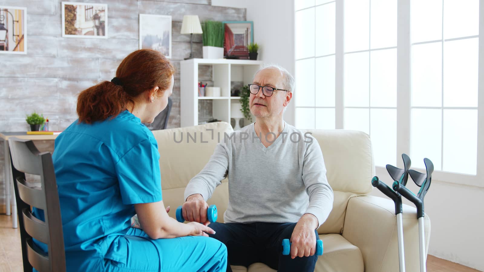 Female nurse helps an old man to make his morning exercises in bright and cozy retirement home