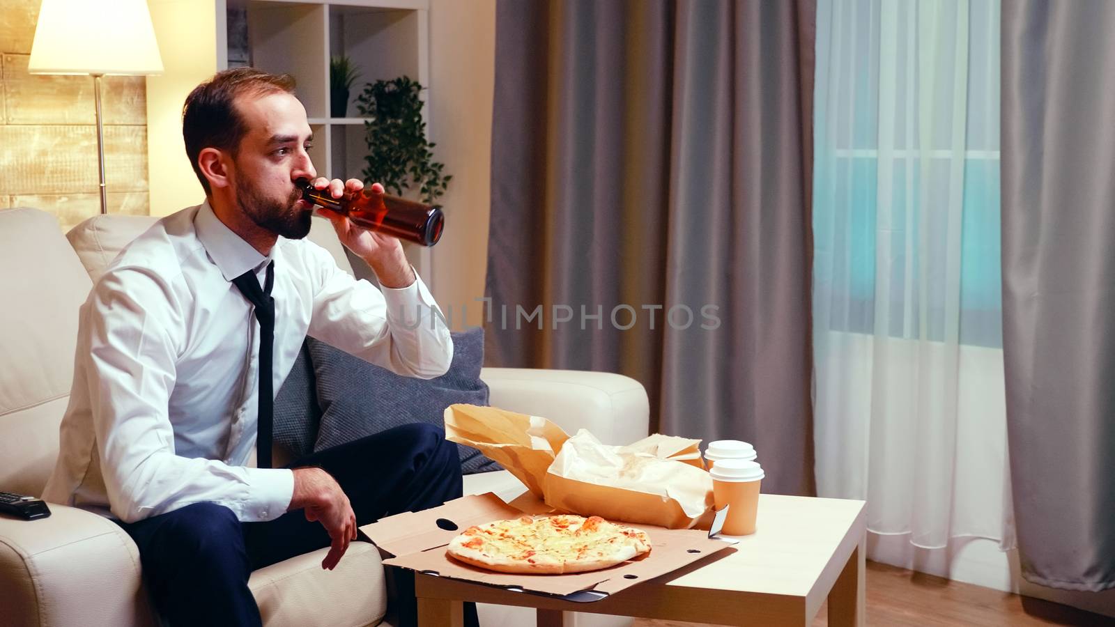 Businessman resting on the couch drinking beer and eating pizza in living room. Relaxing after work watching tv.