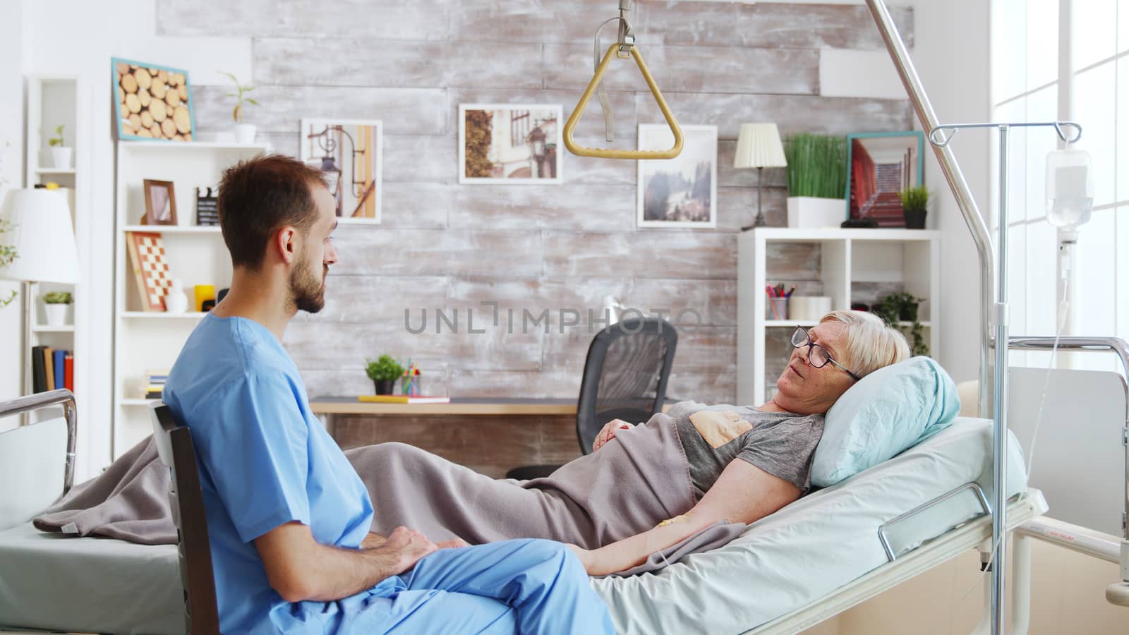 Male assistant taking a seat near hospital bed of ill lady lying in nursing home with big and bright windows.