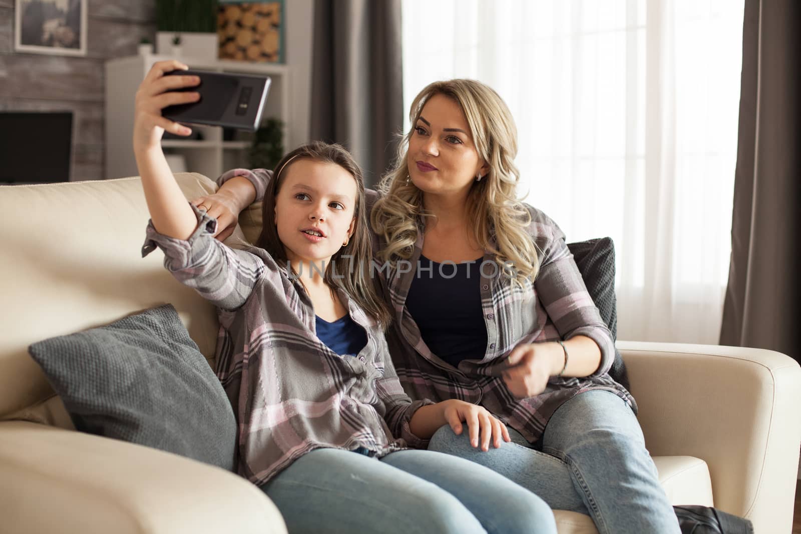 Mother and little daughter sitting on the couch in living room taking a selfie.