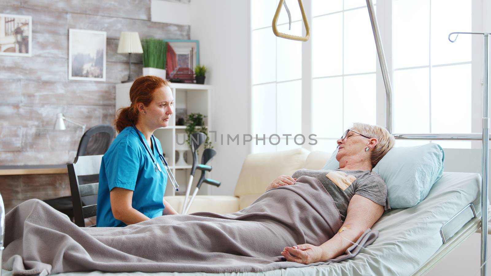 Nurse in retirement home talking with an old lady lying in hospital bed. Big windows with bright light are behind