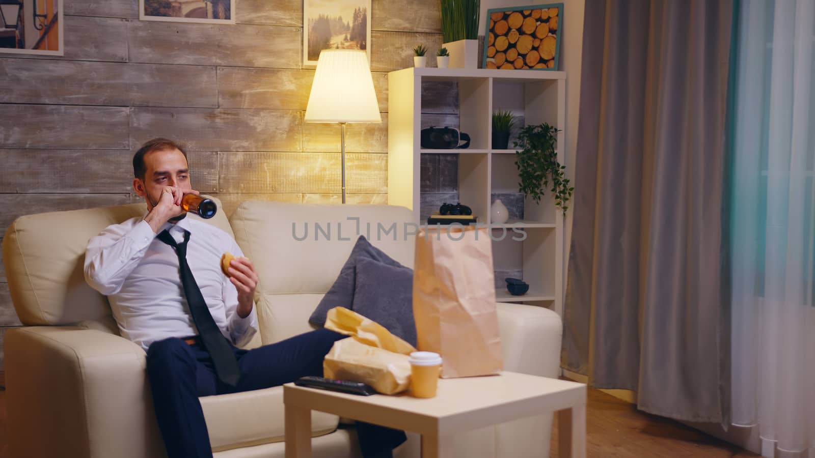 Starving businessman in suit after a long day at work eating burger and using tv remote control. Unhealthy food.