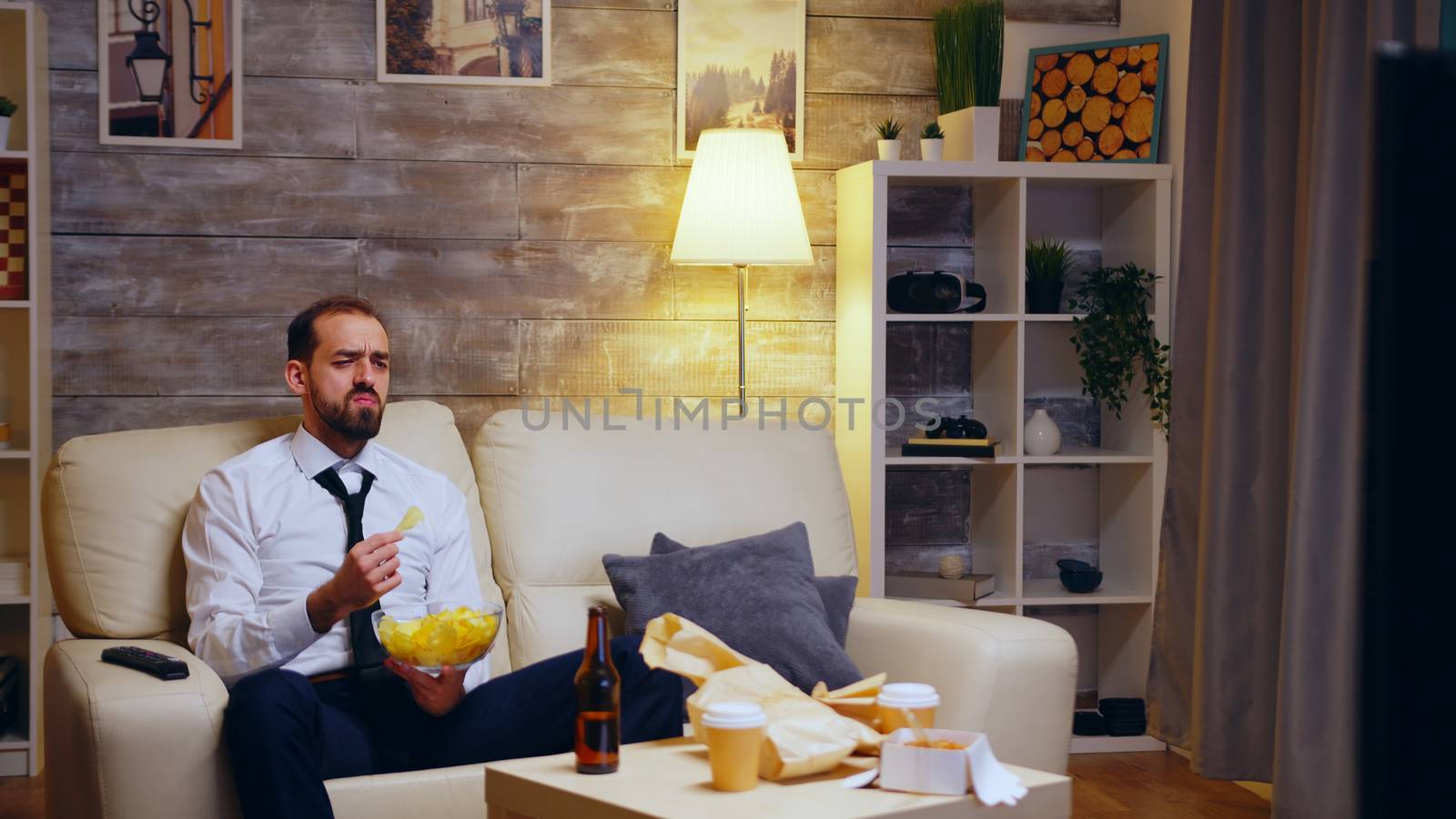 Businessman sitting on couch eating chips eating television. Zoom in shot.