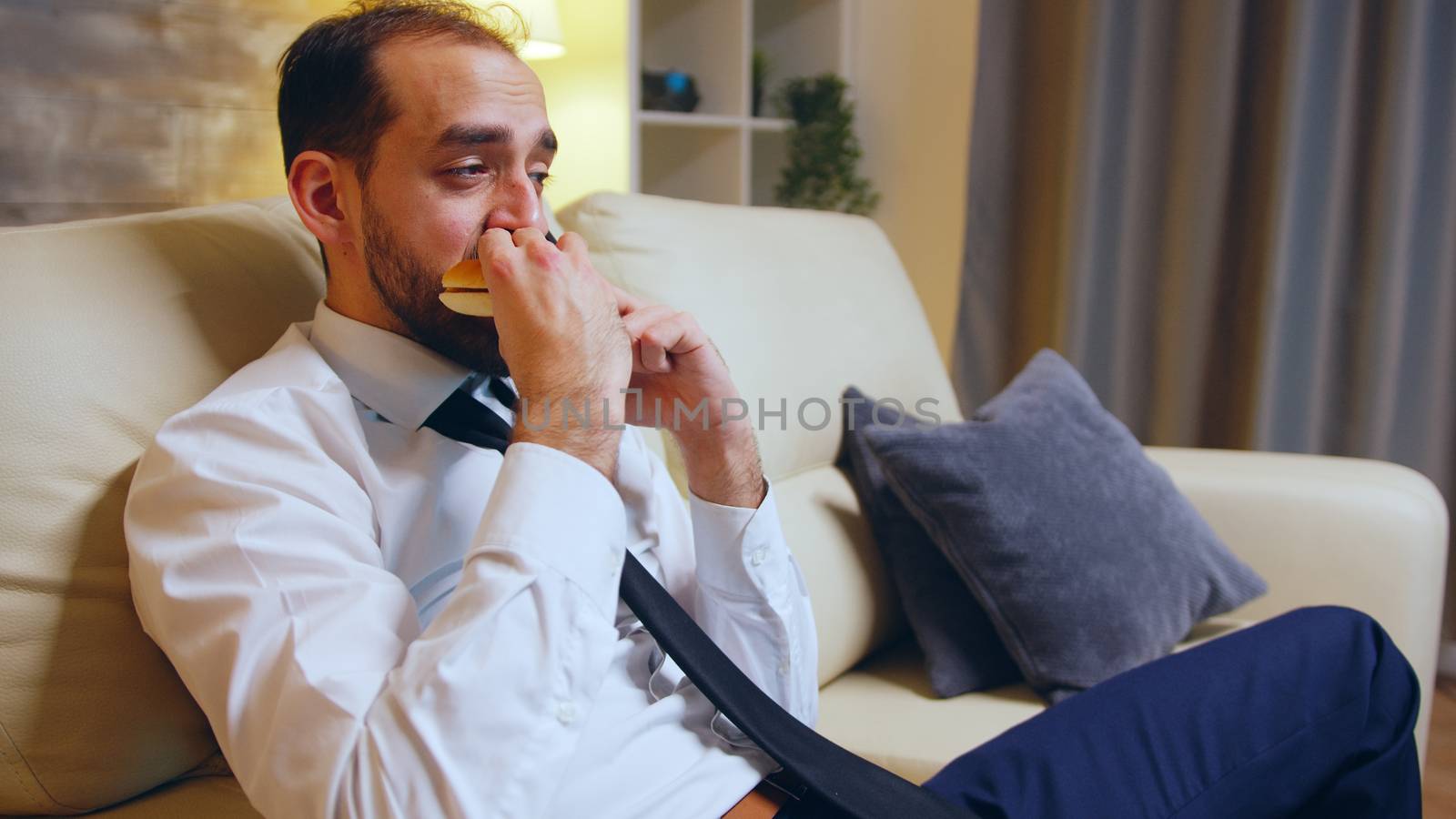 Businessman in formal wear sitting on couch eating a burger and talking on the phones after a tiring day.