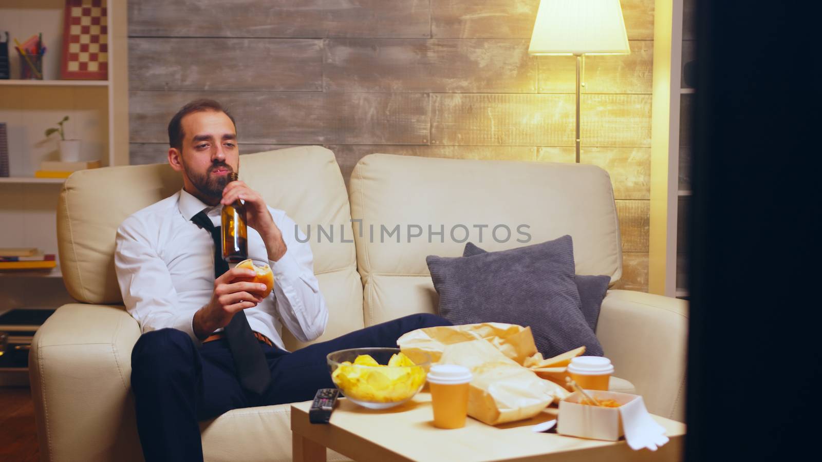 Businessman with tie sitting on couch eating a burger and talking on the phone.