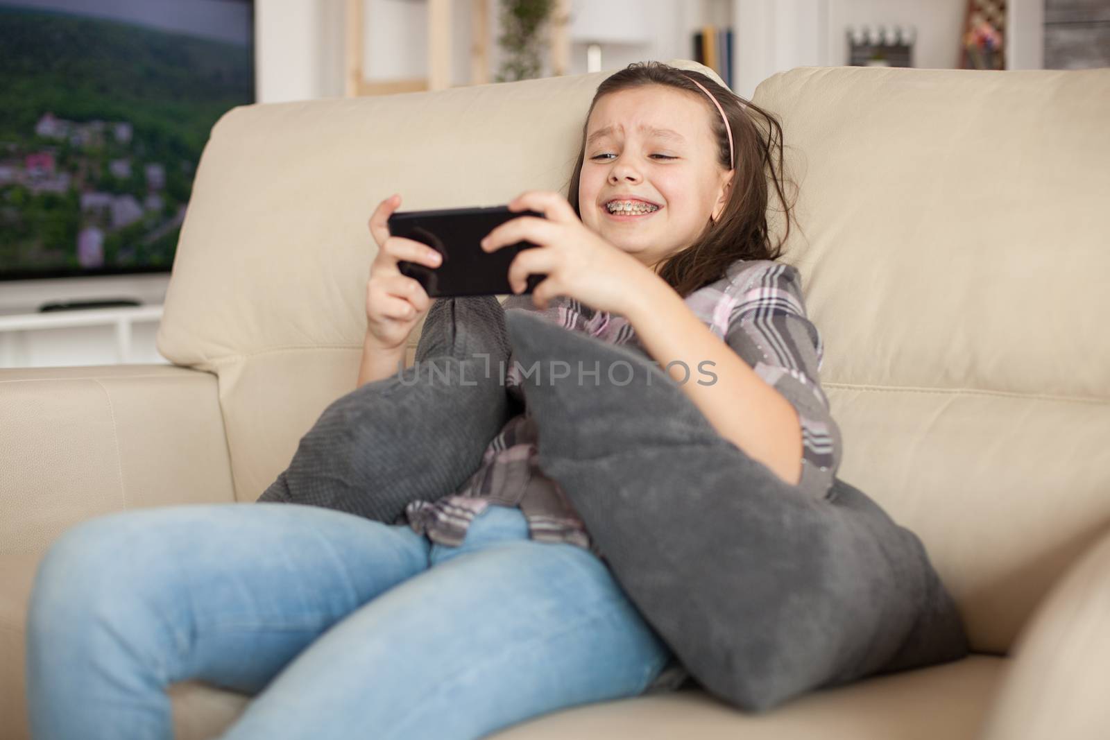 Cheerful little girl playing video games on smartphone lying on the couch.