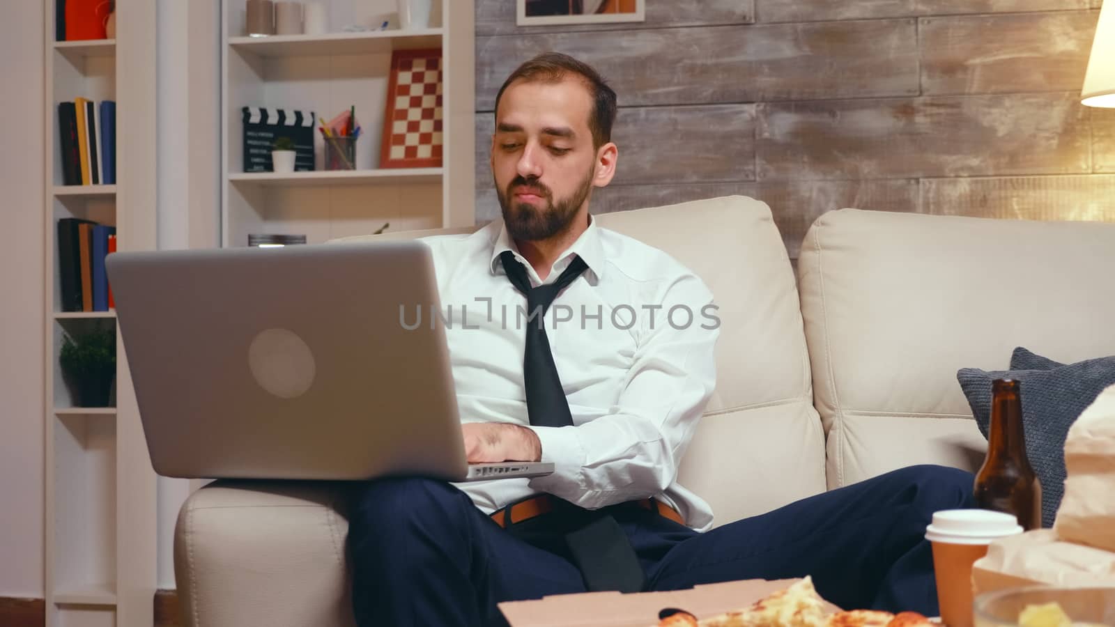 Tired businessman in living room working on laptop and eating pizza. Entrepreneur wearing a tie.