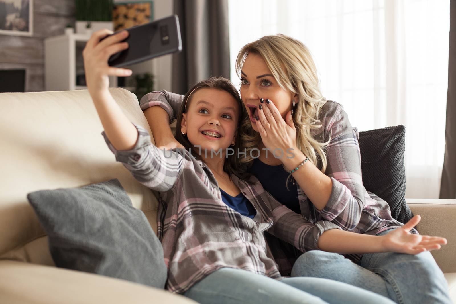 Cheerful kid and her mother are taking a selfie in living room sitting on the couch having a good time.