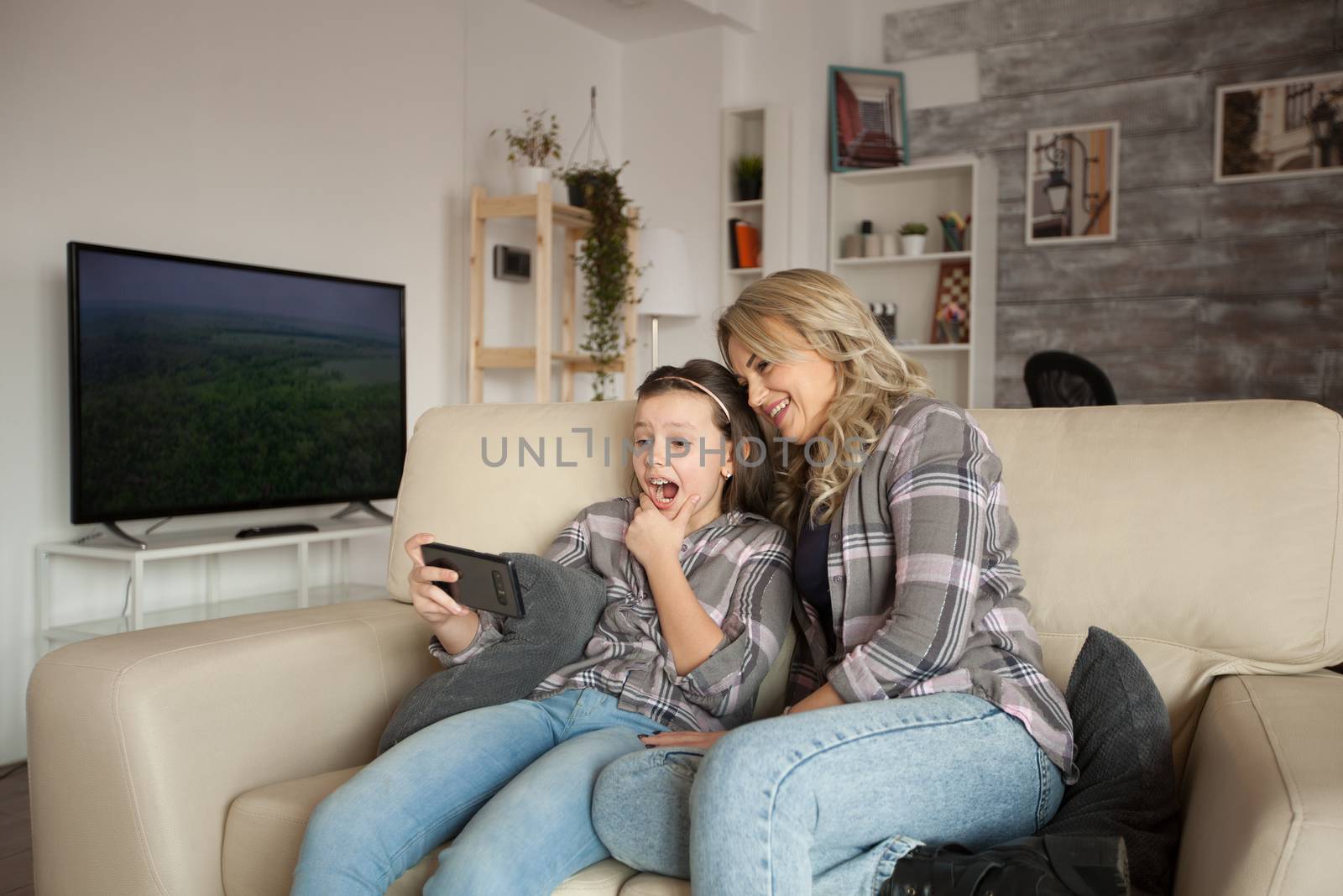 Shocked little girl white braces while using her smartphone sitting next to her mum on the couch in living room.