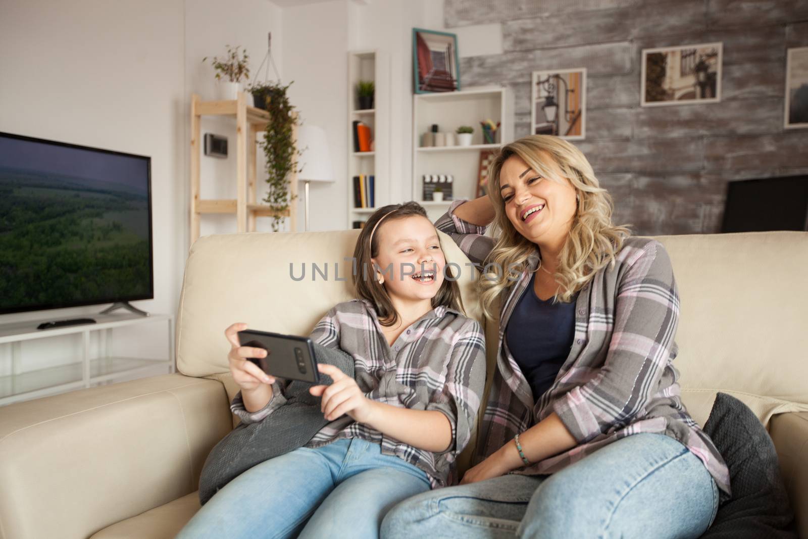 Little girl with braces smiling and pointing at phone screen sitting with her mother on the couch.