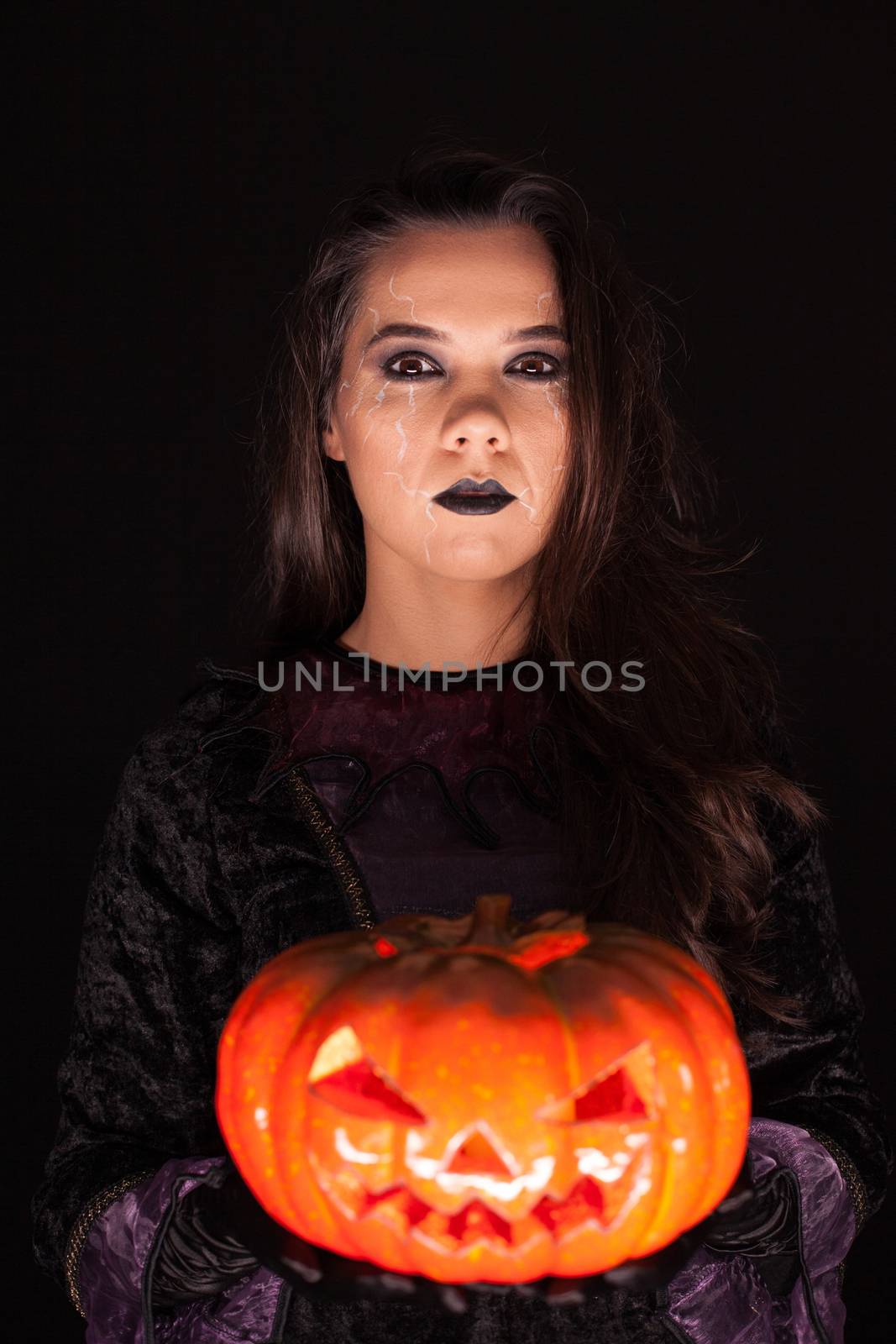 Attractive woman with a serious face dressed up like a witch holding a pumpkin over black background.