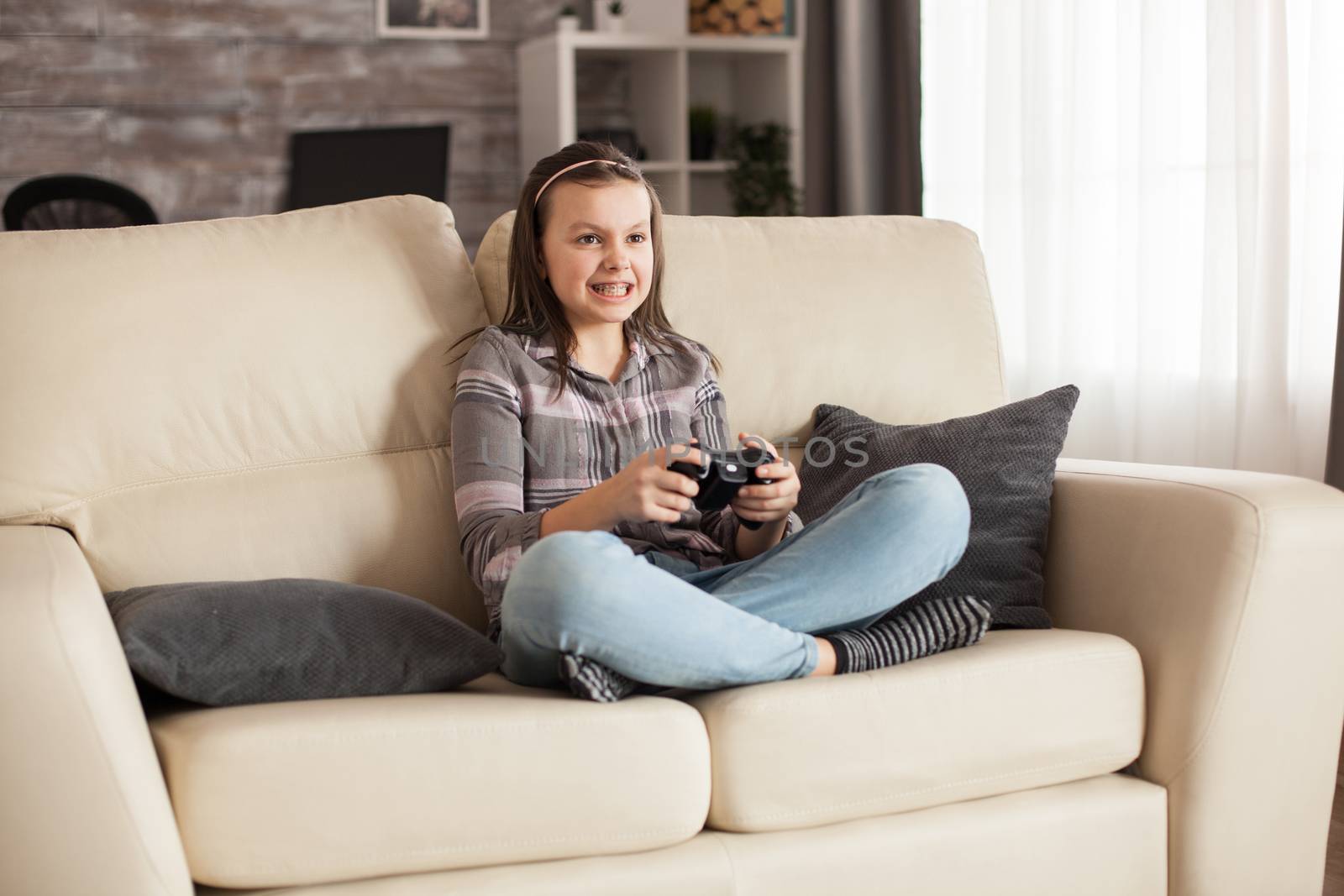 Little girl with braces sits on sofa playing video games by DCStudio