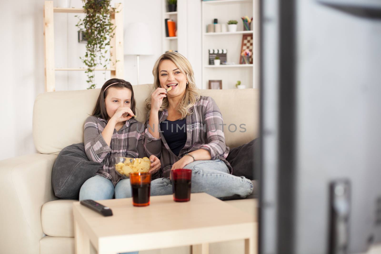 Young mother and her teenage girl watching a film on tv sitting on sofa eating chips.