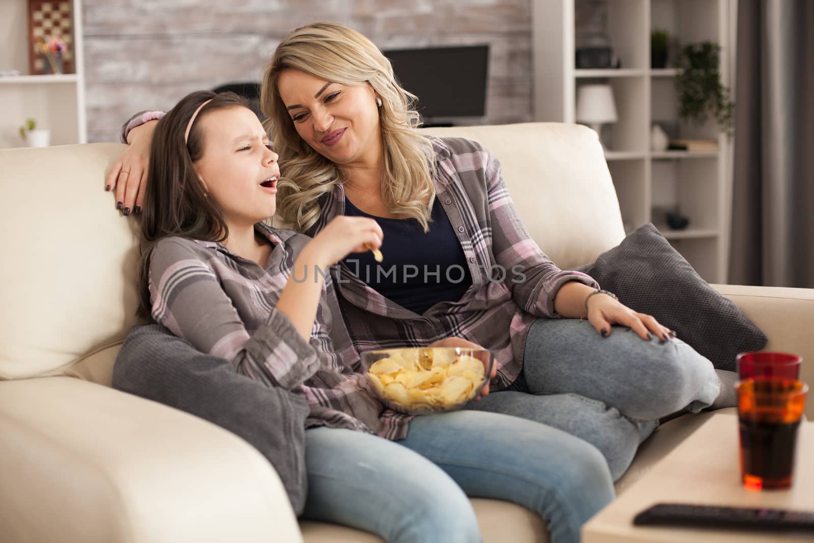Young mother looking at her teenage daughter while sitting on sofa and watching tv. Happy little girl.