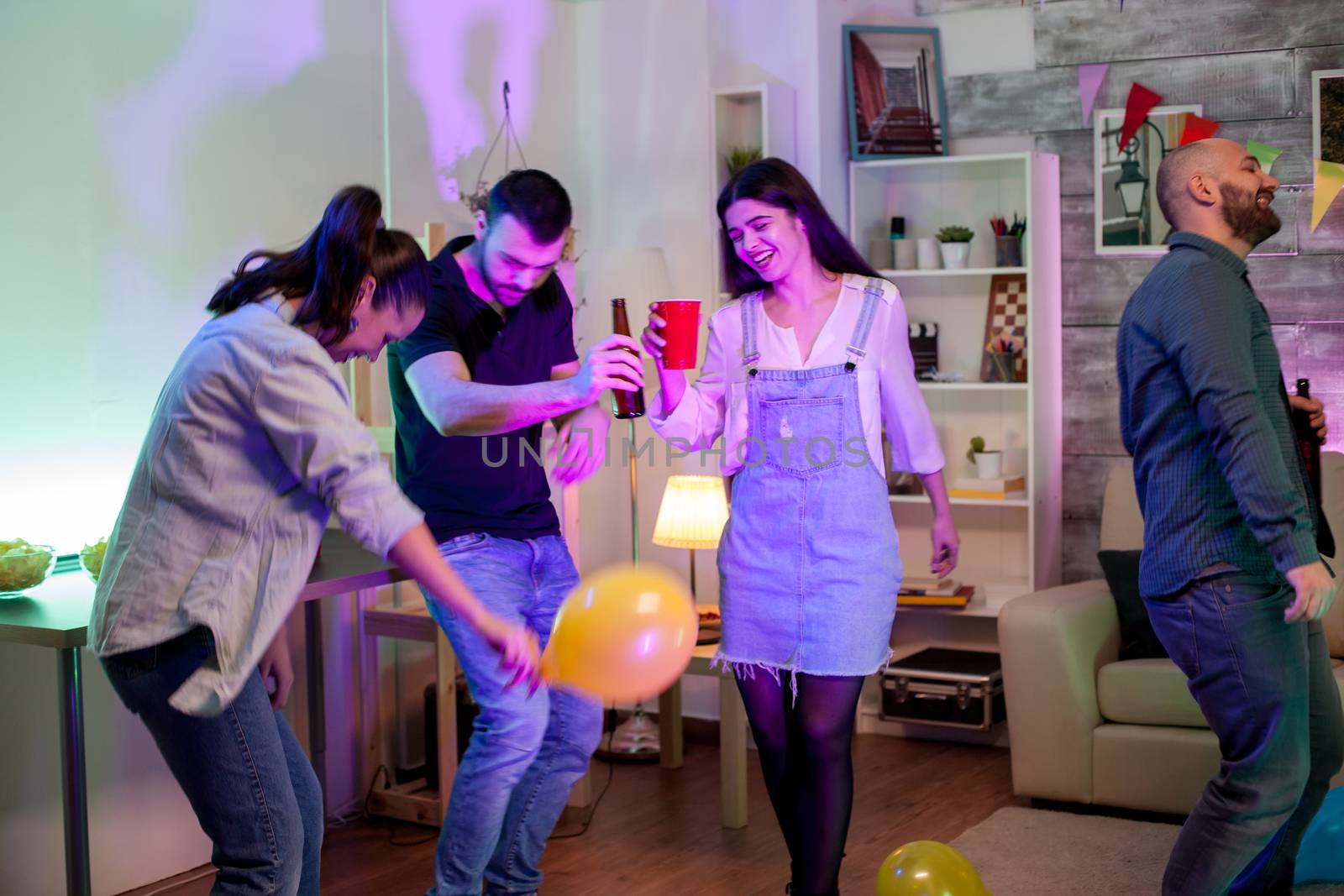 Cheerful young man holding a beer bottle while dancing with his friends at a party with disco music and balloons.