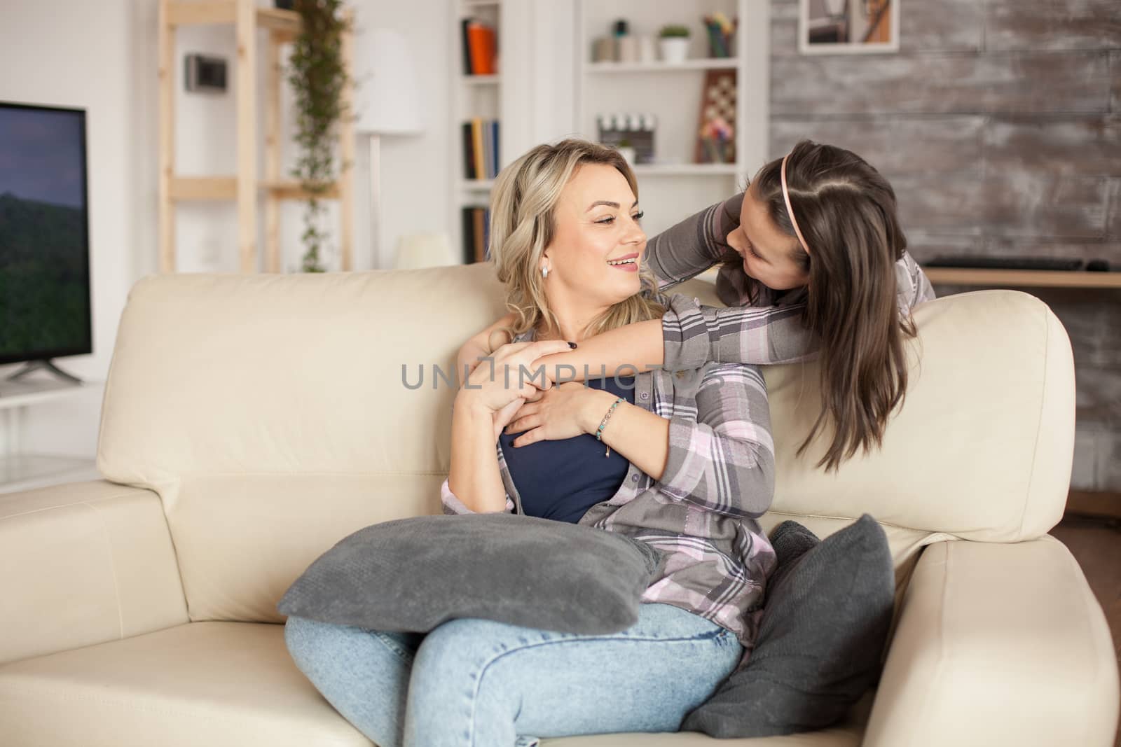 Young mother sitting on the couch hugged from behind by cheerful little daughter.