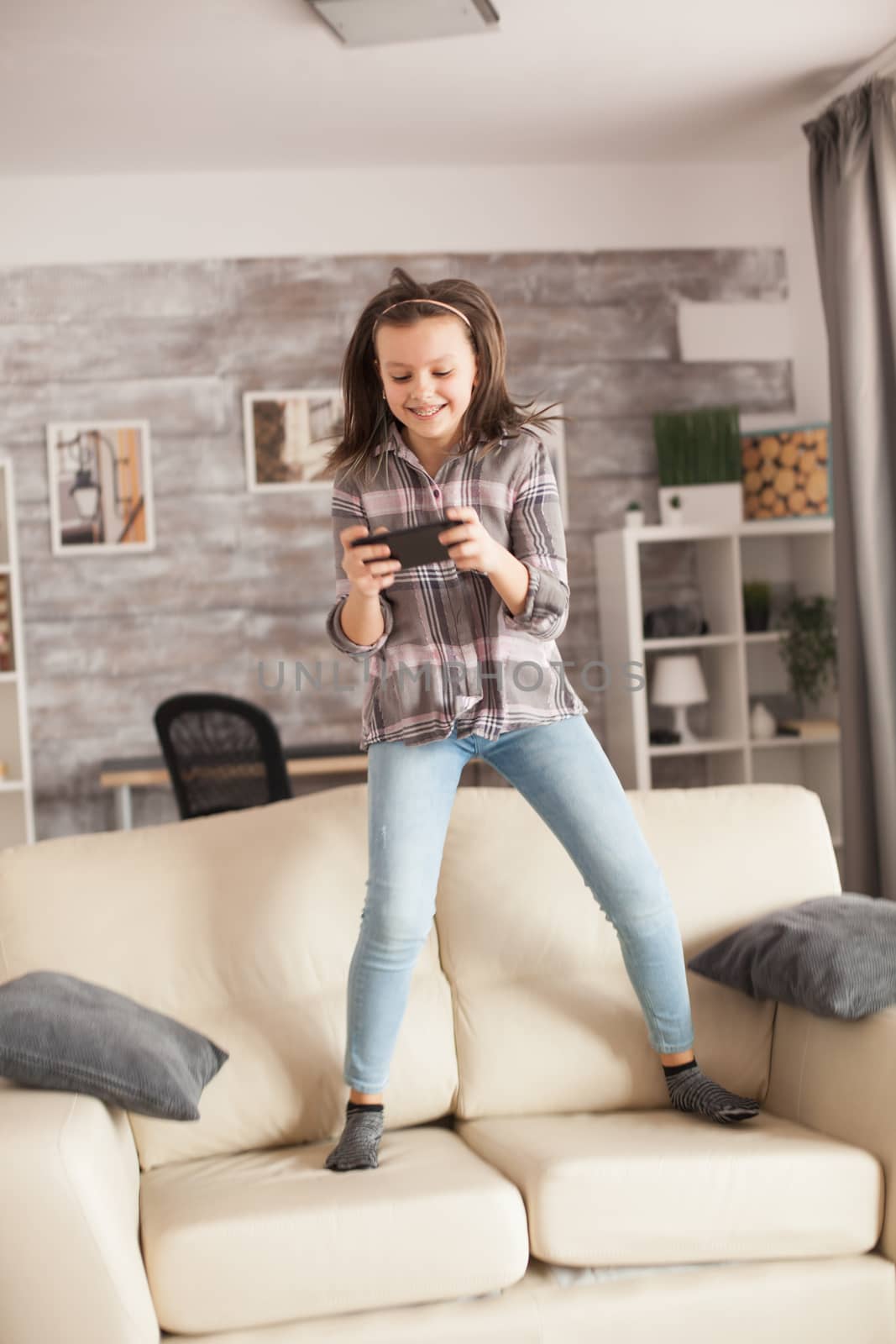 Excited little girl using her smartphone and jumping on the sofa.