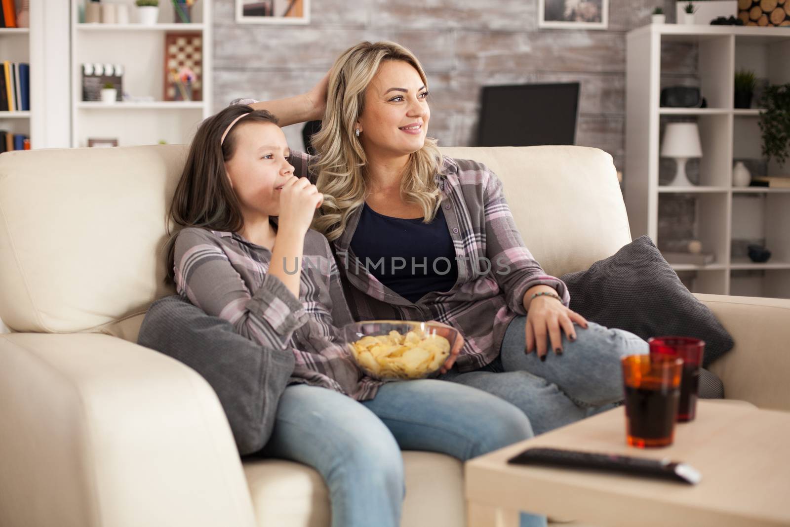 Relaxed young mother and cheerful daughter watching tv sitting on sofa eating chips.