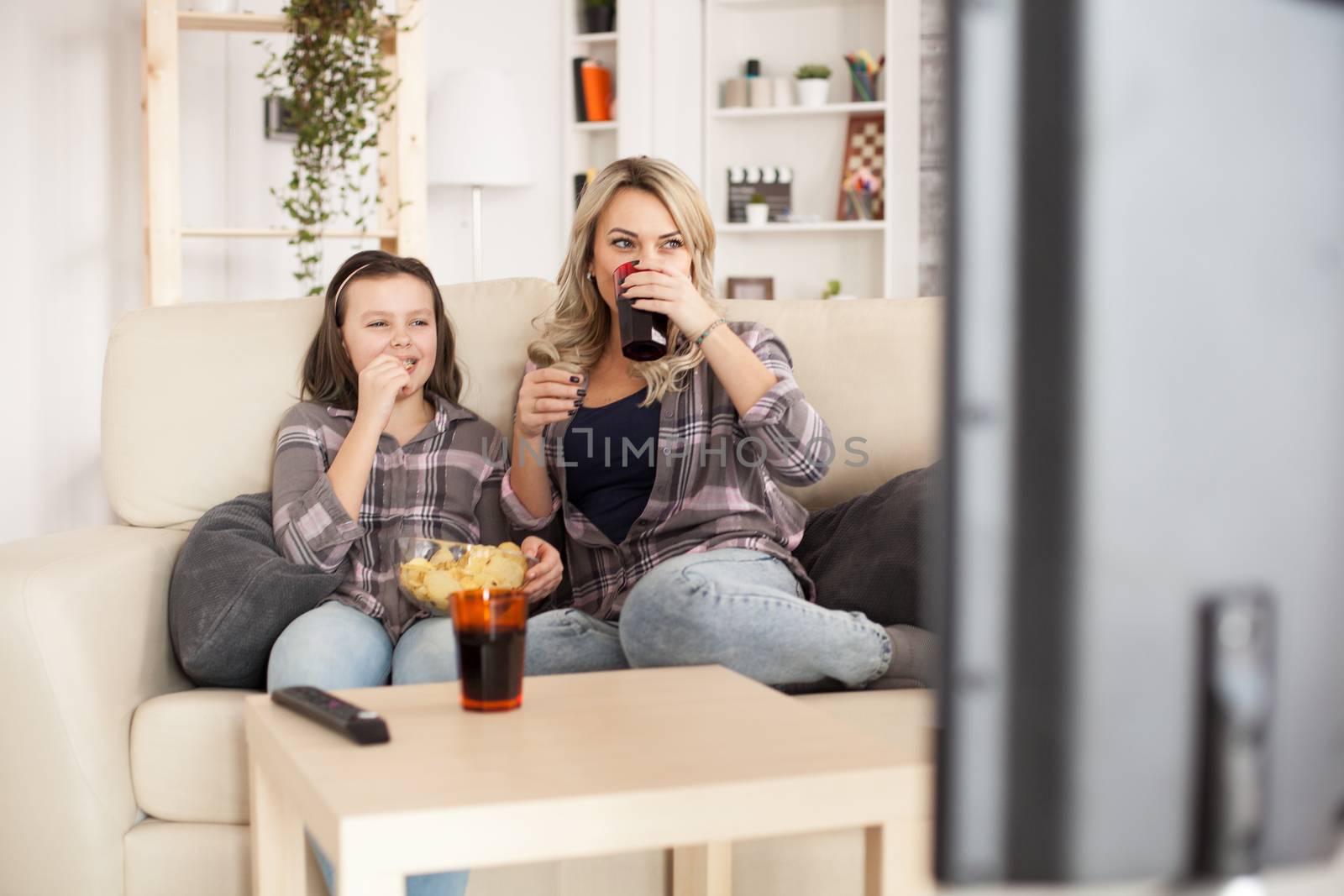 Happy young mother sits on couch with her teenage daughter watching a film on big lcd.